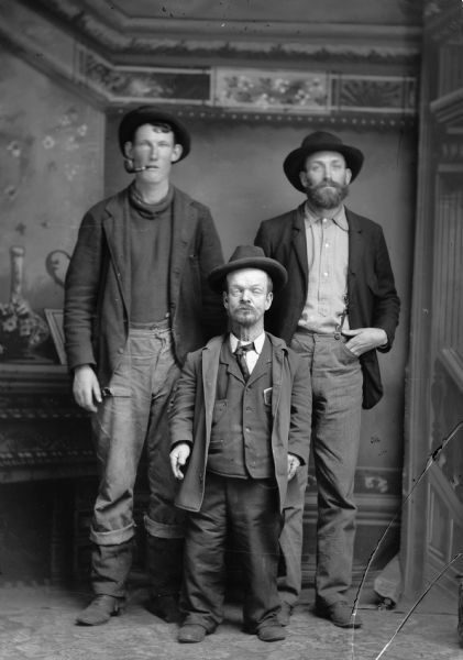 Studio portrait of three men standing, one of whom is a little person, standing in front of a painted backdrop. All of the men are wearing hats, and the man on the left has a pipe in his mouth.