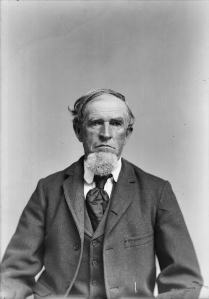 Studio portrait of a seated man with a beard wearing a suit jacket, vest, and necktie.