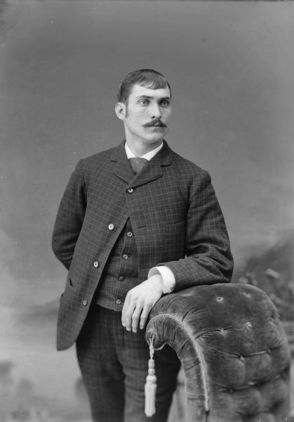 Studio portrait of a man with a moustache standing and leaning on a stuffed chair in front of a painted backdrop. He is wearing a suit jacket with matching trousers, vest, and tie.