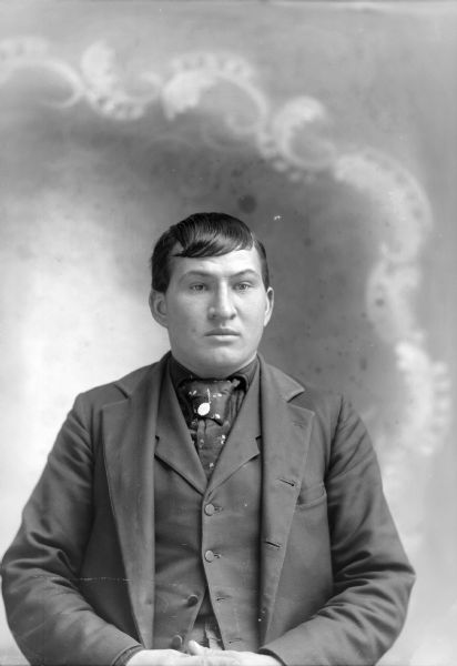 Studio portrait in front of a painted backdrop of a seated young man wearing a suit jacket, necktie, and vest. There is a tie pin or charm attached to his necktie.