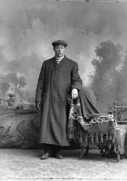 Studio portrait of a standing man in a long overcoat, wearing a hat and leaning on a chair which is covered with a tasseled blanket. He is holding one of his gloves and is posing in front of a painted backdrop.
