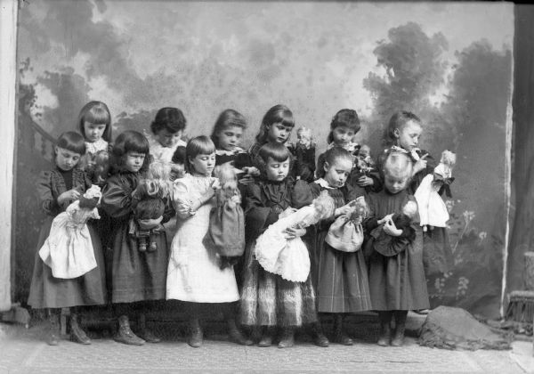 Studio portrait of a dozen young girls standing and looking down at dolls they are holding in their left hands. They are all wearing dresses and pointing at their dolls. They are posed in front of a painted backdrop.