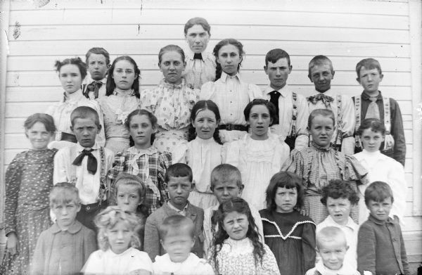 A woman and a group of children posed standing in front of a wooden building, possibly a grammar school class photograph. The girls are wearing dresses, and the older boys are wearing neckties.