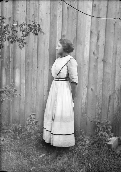 Full-length portrait of a woman wearing a long dress, posed standing in the grass in front of a wooden wall.