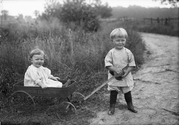 An outdoor portrait of a small child posed sitting in an "Iron Clad" coaster wagon pulled by another child. They are standing outdoors on a road or path, with a field and fence in the background.
