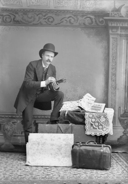 A studio portrait of a man who is posing standing with one leg resting on luggage and holding a pencil and a bill pad in front of a painted backdrop. He is wearing a hat, a long jacket over a vest, and a necktie. There is a watch fob attached to his vest. Possibly a traveling salesman. On the table near the man is a book open with a display of fasteners and suspenders, perhaps for clothing. Suitcases and valises are on the rug in front of him.