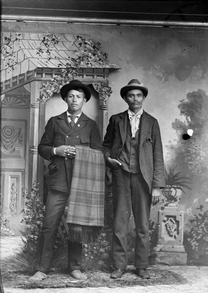 Studio portrait of two Ho-Chunk men posing standing in front of a painted backdrop. The man on the left holding the blanket, is Louis Bearchief. Both men are wearing hats and neckties, with suit jackets and trousers.