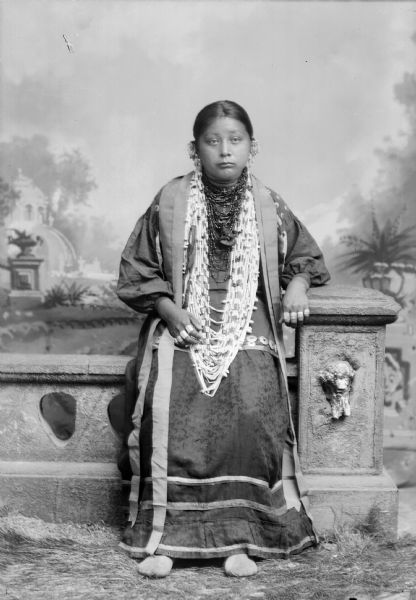 Full-length studio portrait of a Ho-Chunk woman. She is sitting on a prop stone wall and is wearing a dress with earrings and long strands of beads. She is wearing rings on eight of her fingers. In the background is a painted backdrop. Her name is Grace Decorra, TwoCrow, Winneshiek, Massey, born in 1877 and died 9/1914, (MaHeHutTaWinKah) (daughter of [MauCooChoEMonEKah] David Decorra and [NauChooPinWinKah] Kate Decorra, Otter) (Grace  was the former wife of Albert TwoCrow and Edward Winneshiek) Her last marriage was to William Massey.