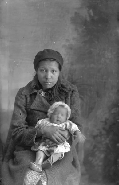 Studio portrait in front of a painted backdrop of a Ho-Chunk woman wearing a hat over shoulder-length hair and wearing a winter coat over a dress. She is sitting and holding in her lap an infant who is wearing a cap, shirt, leggings, and moccasins. Probably Marie Big Hawk Whitewater.