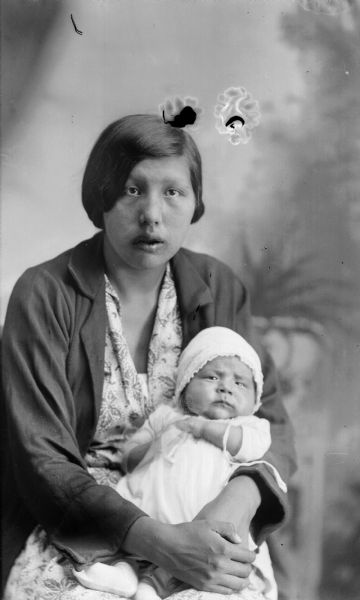 Studio portrait in front of a painted backdrop of a Ho-Chunk woman with short hair, wearing a jacket, and dress. She is posing sitting and holding in her lap a child wearing a long gown, a cap, and booties. Probably Amanda Big Hawk Stacy holding Irving Little George.