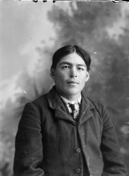 Waist-up studio portrait of a Ho-Chunk man posing sitting. He is wearing a suit and tie. Probably Charley Blackdeer.