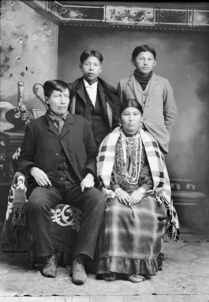 Full-length studio group portrait in front of a painted backdrop of a Ho-Chunk man and woman posing sitting in front of two Ho-Chunk boys. The men are wearing suit jackets, vests, and trousers. The woman is wearing a long dress, earrings, necklaces, bracelets, and a shawl over her shoulders. Probably Mr. and Mrs. John Blackhawk and sons.