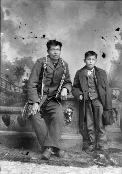 Full-length studio portrait in front of a painted backdrop of two Ho-Chunk males posing sitting and leaning against a prop stone wall. Probably John Blackhawk on the left, wearing a wool sash on his shoulders over his coat, and Albert Blackhawk on the right, holding a hat and wearing a coat, suit jacket, vest with watch fob, and trousers.