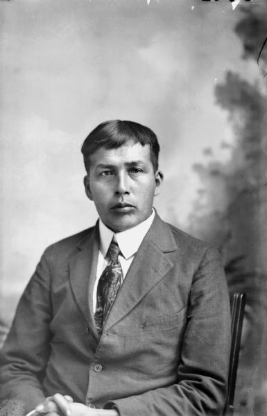 Waist-up studio portrait in front of a painted backdrop of a young Ho-Chunk man posing sitting. He is wearing a suit jacket and necktie. Probably John Blackhawk.