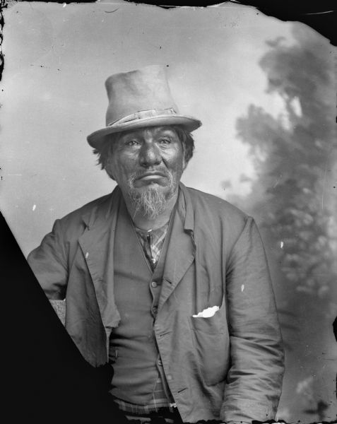 Waist-up studio portrait in front of a painted backdrop of an elderly Ho-Chunk man with a beard. He is wearing a suit jacket, vest over a plaid shirt, and a hat. Probably Little Blackhawk.