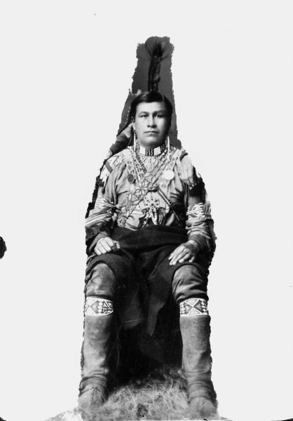 Full-length studio portrait of a Ho-Chunk man in regalia. The background around him has been whited out. He is wearing a beaded collar, earrings, a decorated shirt, and trousers with leggings. Probably Little Blackhawk.