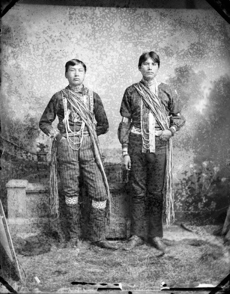 Full-length studio portrait of two young Ho-Chunk men standing and wearing regalia, including wampum strings. Behind them is a prop stone wall. Standing (l to r) Frank Redcloud and Jasper Blowsnake (WaReTcaWeRa). In the background is a painted backdrop.