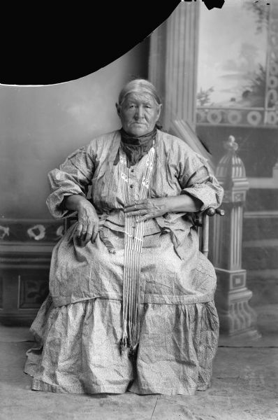 Full-length studio portrait of an elderly Ho-Chunk woman sitting and wearing long bead strands. In the background is a painted backdrop. Mary Snowball, Decorra, Buffalohead (PayDokSeKah [Edge of brow is yellow]), born 1863 and died November 20th, 1940. She was the wife of John Buffalo Head (ChayPayKay) and the mother of five children: Grace, John, Stella, Frank and Carrie. (Daughter of Snow Ball [WauSooMonEKah] and [WaConChaWinKah]) (Maternal granddaughter of Iron Walker and Green Feather Woman). 
