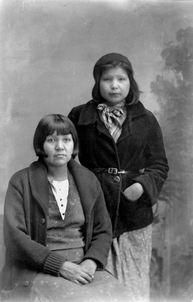 Three-quarter length studio portrait of two Ho-Chunk women, with short hair, posing in front of a painted backdrop. The woman on the left is sitting, and is wearing a sweater over her dress, and the other is woman is standing and wearing a short, belted coat over a dress, a scarf around her neck, and a hat. Agnes Payer Climer is on the left, and Bertha Climer is on the right.