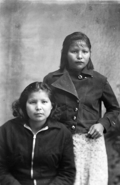 Studio portrait of two Ho-Chunk women in short hair in front of a painted backdrop. One is posed sitting wearing a zippered jacket. The other woman is standing on the right and is wearing a short, buttoned jacket over a skirt or dress. Agnes Payer Climer is on the left, and Bertha Climer on the right.