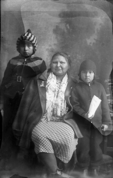 Studio portrait of a Ho-Chunk woman posing sitting in the center, flanked by two children, in front of a painted backdrop. They are all wearing modern winter dress. Agnes Climer is with Juanita Climer, wearing a striped hat, on the left, and John Climer, on the right, wearing a cap and holding a bag in his hand.