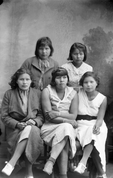 Studio portrait of five young Ho-Chunk women in front of a painted backdrop. Two are posing standing in the back row, and three are sitting. They are all in modern dress and short hair. Standing from left to right is Jean Climer and Edna Climer, sitting from left to right is Susie Climer, Agnes Payer Climer, and Bertha Climer; all the women are daughters of Frank Climer.