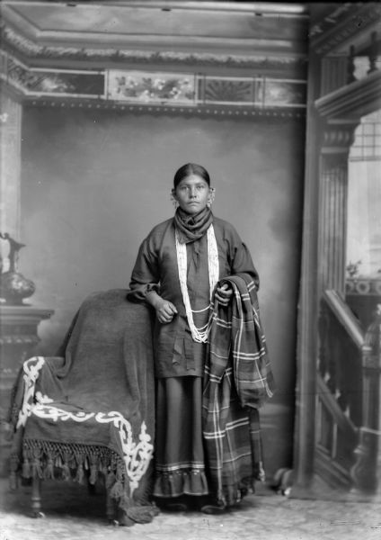 Studio portrait of a young Ho-Chunk woman posing standing in front of a painted backdrop near a chair. She is holding a wool shawl, and is wearing earrings. She is also wearing a bird bone necklace. Probably the wife of John Davis.