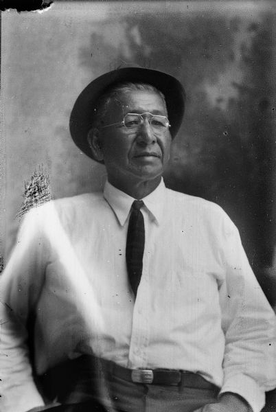 Studio portrait of John Davis posing sitting and wearing a white shirt, tie, eyeglasses, and a hat. He is sitting in front of a painted backdrop.