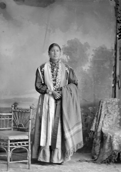 Studio portrait of a young Ho-Chunk woman, Grace Decorah Whitegull, posing standing next to a chair in front of a painted backdrop. She is holding a wool shawl, and is wearing a dress, earrings, bracelets, rings, and white bead necklaces.