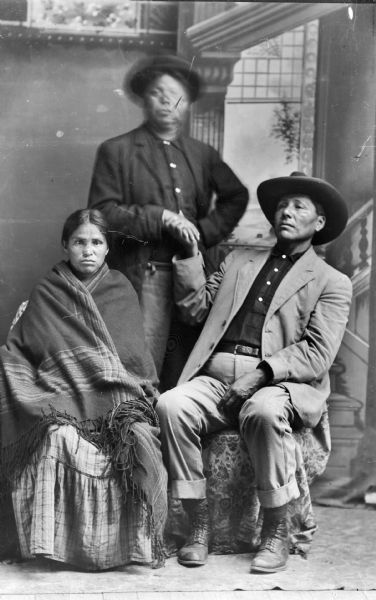 Studio portrait in front of a painted backdrop of two Ho-Chunk men in modern dress and wearing hats, and a woman wearing a wool shawl over a dress. The man standing is blurred by movement, and he is shaking hands with the man sitting on the right. The woman is probably the wife of Will Decorah.