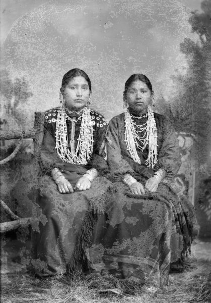 Studio portrait of two young Ho-Chunk women posing sitting in front of a painted backdrop and a studio prop fence. They are wearing white bead necklaces and bracelets, and are holding wool shawls on their laps. The woman on the left is the daughter of Mary Eagle.