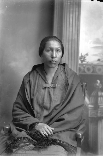 Studio portrait of a Ho-Chunk woman sitting in a chair wearing a black dress and with a wool shawl wrapped around her shoulders. Probably Flora Eagle, the daughter of George Eagle. She is posing in front of a painted backdrop.