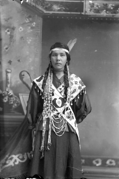 Studio portrait of a Ho-Chunk woman, Nellie Eagle Swallow, posing standing and wearing very long Winnebago earrings, a beaded headband with two eagle feathers, and bandoleers. She is posing in front of a painted backdrop.