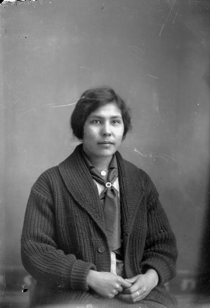 Studio portrait of a Ho-Chunk woman posing sitting in front of a painted backdrop. She is wearing a dark sweater, a necklace, and a scarf with a pin. Probably Nellie Eagle, the wife of Tom Wallace.