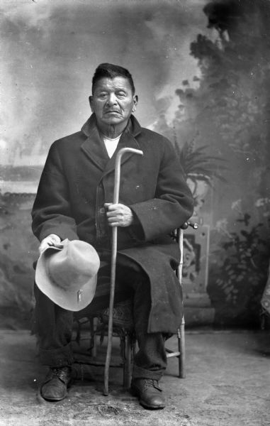 Studio portrait of an elderly Ho-Chunk man in a coat and trousers posing sitting, holding his cane and hat in front of a painted backdrop. He is James Eagle, the custodian of the Decorah (French) war bundle.