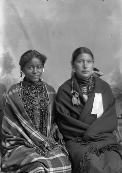 A studio portrait of two young Ho-Chunk women posing sitting in front of a painted backdrop. They are wearing beaded necklaces, long earrings, and are wrapped in shawls. Carrie Elk on the left, is of African and Native American heritage.