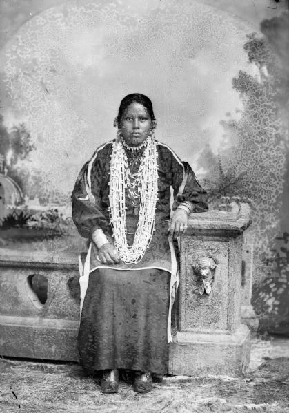 Studio portrait of a Ho-Chunk woman posed sitting on a prop stone wall in front of a painted backdrop. She is wearing a white bead necklace, bracelets, rings, and earrings. Probably Annie Falcon, later the wife of William Hall, Sr.