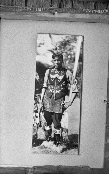 Copy photograph displayed on a board of a Ho-Chunk man posing standing outdoors and wearing full regalia. Probably Harold Jones Funmaker, also known as Wanajiska (trans. Fastest Man in Race).
