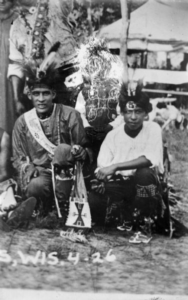 Copy photograph of two Ho-Chunk men posing kneeling, and a third boy standing, but etched out of the negative. They are all wearing full regalia. The man on left is Harold Jones Funmaker, also known as Wanajiska (trans. Fastest Man in Race). There is a tent in the background.