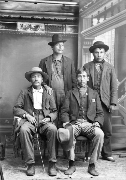 Studio portrait of four Ho-Chunk men, two posing sitting and two standing in front a painted backdrop. The man standing on the right is probably Ed Funmaker. They are all wearing modern dress, including suit jackets, trousers, and overalls. They are wearing or holding hats, and the man on the right is wearing a beaded necklace.