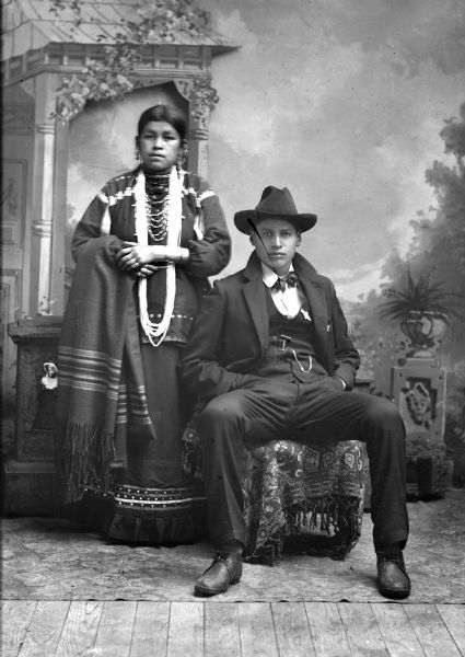 Studio portrait of Will Thunder (NaKikSayWaHeKah) (Son of [WaConChaKah] John Thunder aka Dr. Thunder and [WeHonPeKaw] Lucy Bear, Thunder.) He is wearing a suit jacket, vest with watch fob, tie, and hat. His wife Belle Hall, Greencrow, stands next to him. She is  wearing a long bead necklace and earrings, and holding a wool shawl. They are posed in front of a painted backdrop and prop stone wall.