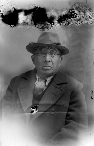 Studio portrait of a Ho-Chunk man, George Greengrass, wearing a suit, tie, hat, and eyeglasses.