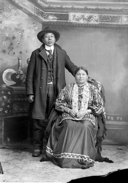 Studio portrait in front of a painted backdrop of a Ho-Chunk man posing standing next to his wife who is posing sitting. They are identified as George Greengrass (WauKeCooPeRayHeKah) and Emma Lookingglass Greengrass (ChayHeHooNooKah).