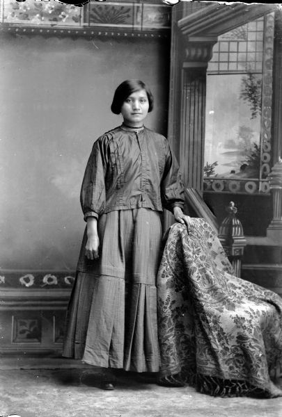 Studio portrait of a young Ho-Chunk woman with short hair posing standing in front of a painted backdrop. She has her hand on a stuffed chair and is wearing a blouse over a long skirt. She is wearing a choker, and a cross on her collar. Probably Ruth Greengrass.