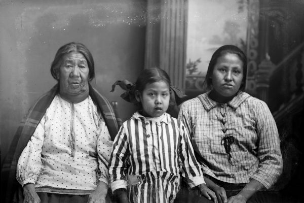 Studio portrait of an elderly Ho-Chunk woman with a shawl over her shoulders posing with two Ho-Chunk girls in front of a painted backdrop. The woman on the left is Fanny Winneshiek, the wife of Grey Eagle, and the girl on the right is Susie Long Marsh.