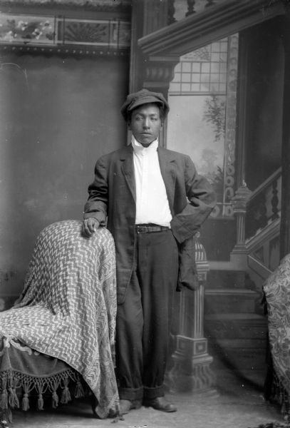 Studio portrait of a Ho-Chunk man in modern dress posed standing in front of a painted backdrop. Probably Willie Goodvillage, who was reportedly blind. He is wearing a suit jacket, shirt, trousers, and a hat, and is resting his arm on a chair covered with a blanket.
