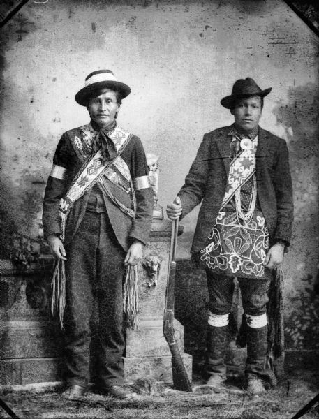 Studio portrait of two Ho-Chunk men standing and wearing hats and regalia. The man on the right is holding a rifle, and the man on the left, William Hindsley, is wearing a bandoleer. They are standing in front of a prop stone wall and a painted backdrop.