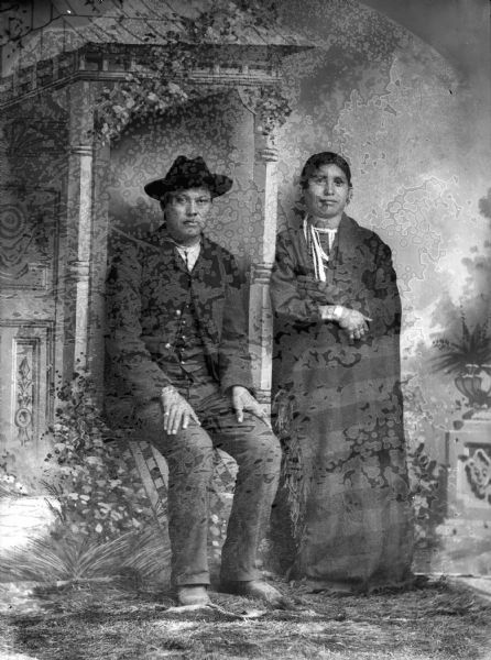 Studio portrait in front of a painted backdrop of a Ho-Chunk man and woman, the former posing half-sitting and wearing a suit jacket, vest, trousers, and a hat, and the latter standing and wearing beads, earrings, and a plaid shawl; the woman is probably Mary Johnson.