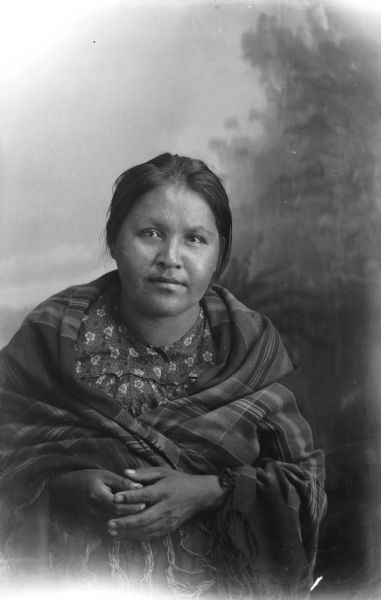 Studio portrait in front of a painted backdrop of a Ho-Chunk woman posing sitting wrapped in a shawl. Probably Minnie Davis, the wife of Frank Davis. She reportedly died in February 1975.