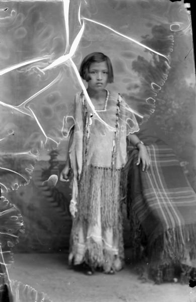 Full-length studio portrait in front of a painted backdrop of Ho-Chunk girl posing standing next to a chair. She is wearing a long buckskin dress with fringe. Probably either Matilda Lewis or Harriet Funmaker.